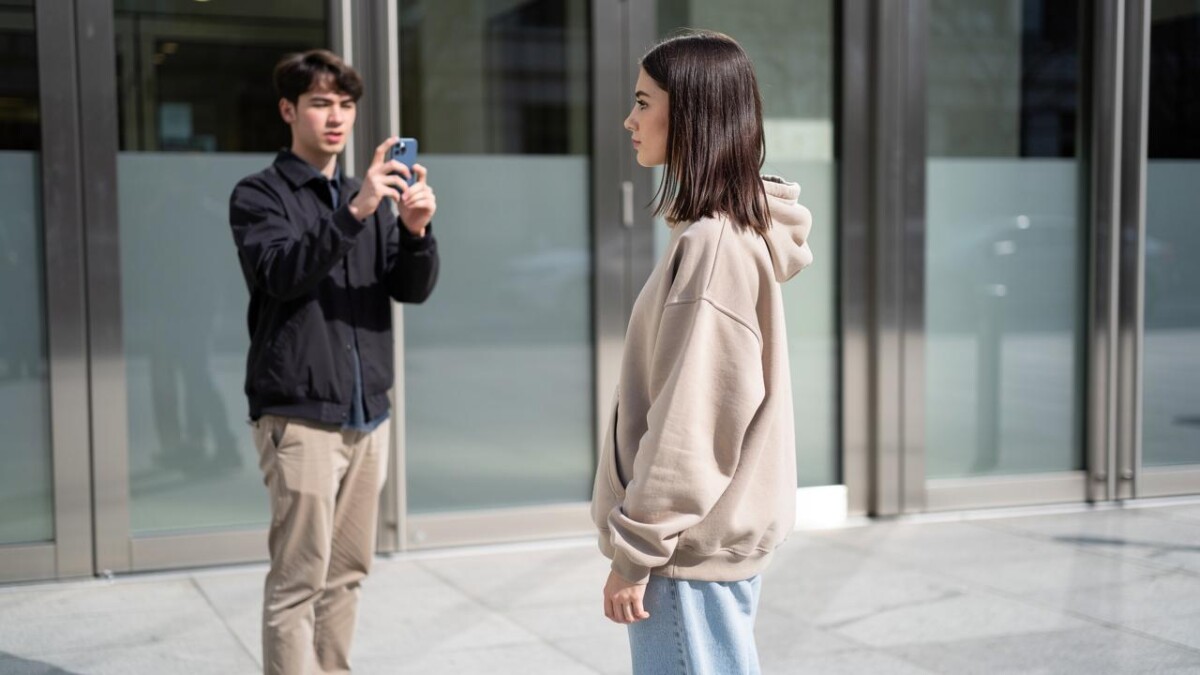 Mlada žena u bež dukserici stoji u profilu dok muškarac fotografiše telefonom